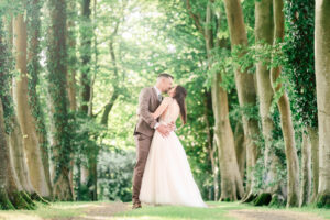 A wedding couple are embracing and sharing a kiss on a tree-lined path in a forest at Crown Lodge, Kent. The groom wears a brown suit, while the bride is in a white, flowing wedding dress. Sunlight filters through the green foliage, creating a romantic, dreamy atmosphere.