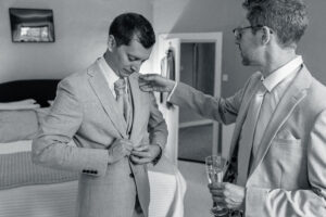 A black and white photo of two men in suits in a bedroom. One man is adjusting his suit jacket while the other is helping him with his tie. The man helping is holding a glass of champagne. The bed and a mirror are visible in the background.