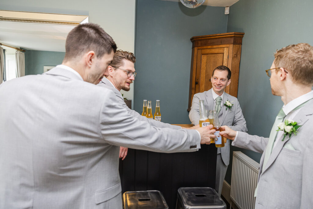 A group of four men in light gray suits are standing in a room, raising their beer bottles for a toast. They are smiling and appear to be enjoying a moment together. One of the men is standing behind a small bar. The room has a wooden cabinet and a radiator.