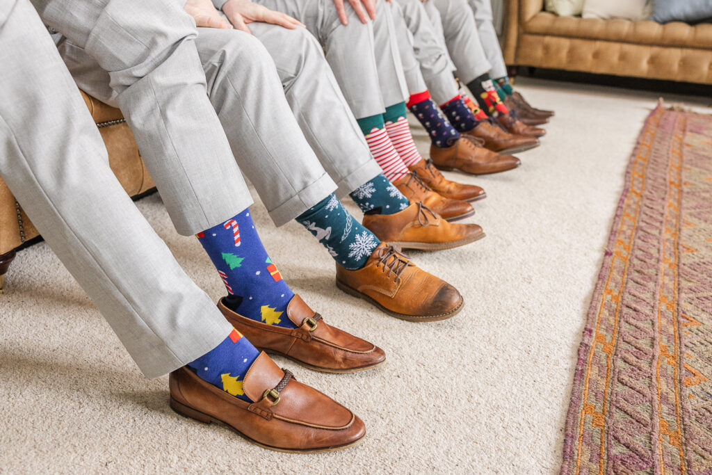 A row of people sit on a sofa, with only their legs visible. They are all wearing light gray pants and brown shoes. Each person is wearing colorful and festive socks with various holiday-themed patterns such as Christmas trees, reindeer, and stripes.