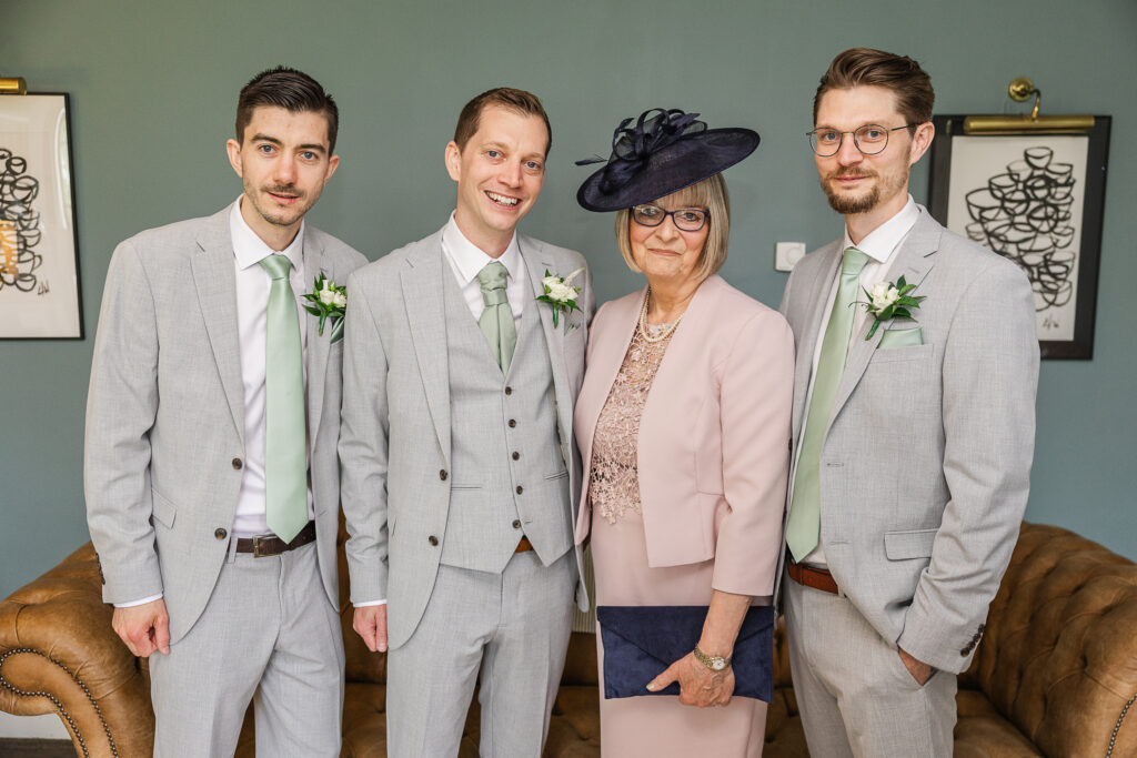 Four individuals, three men in matching grey suits and mint green ties, and one older woman in a pale pink outfit with a matching hat and navy clutch, standing in a living room with dark green walls and framed abstract art. They are all smiling at the camera.