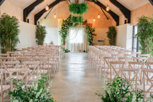 A wedding ceremony setup in a rustic hall, featuring rows of light wooden chairs facing an altar adorned with lush greenery and white flowers. The ceiling has hanging foliage and warm industrial-style pendant lights. The room has a cozy, inviting atmosphere.