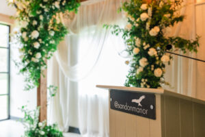 A decorated wedding arch with draped white fabric and white flowers stands in the background of the ceremony space. In the foreground, a podium with a microphone has a sign featuring an elegant bird graphic and the text "@sandomanor.