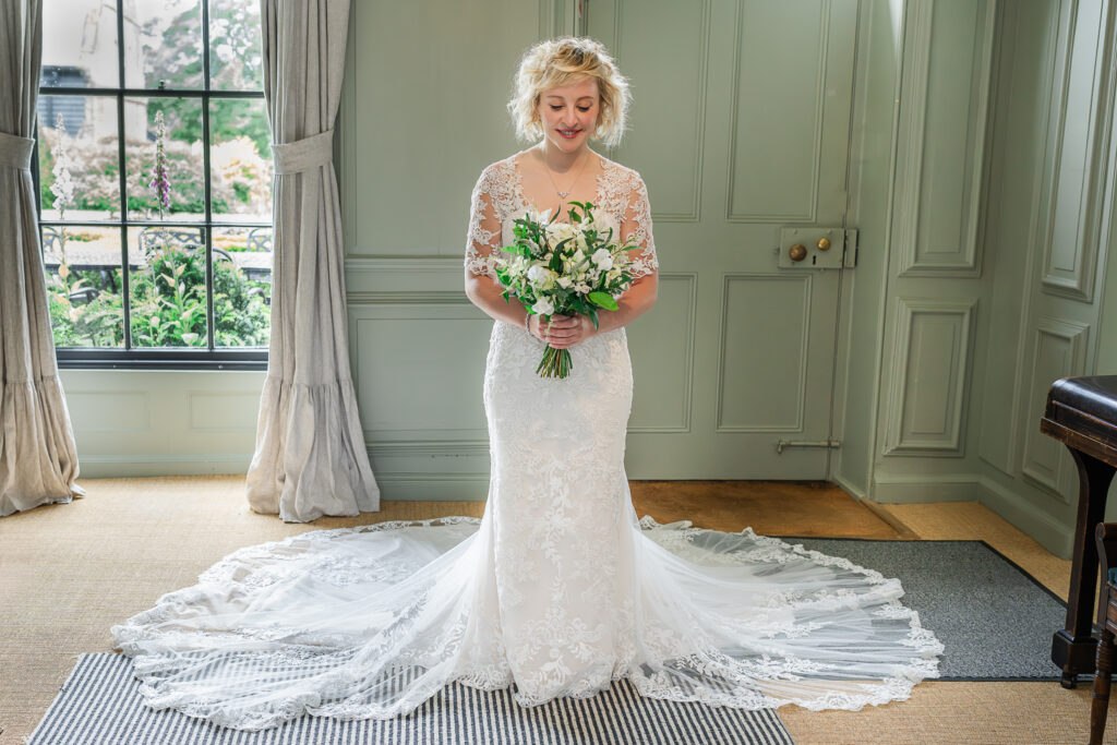 A bride with curly blonde hair stands indoors wearing a white lace wedding dress with a long train. She holds a bouquet of white and green flowers, looking down and smiling softly. The room has light green walls, tall windows with curtains, and lush greenery outside.