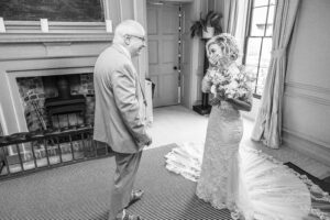 A bride in an intricate lace dress and veil covers her mouth, smiling, as she holds a bouquet and stands near an older man in a suit. They are indoors next to a fireplace and window, with the older man looking at her with a joyful expression.