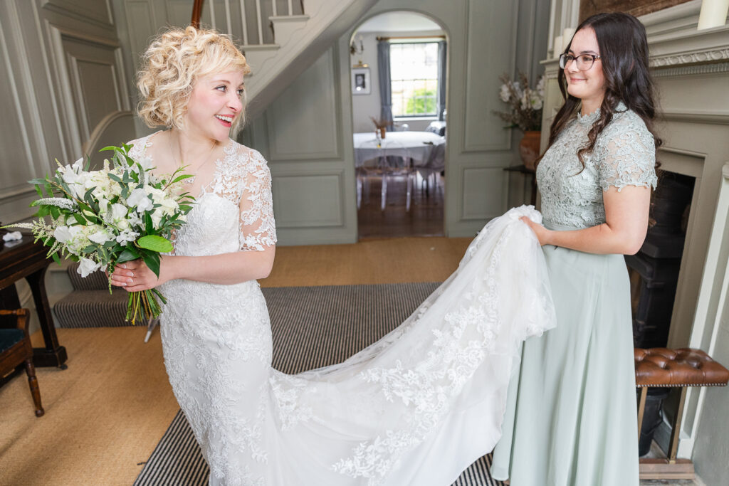 A bride in a white lace wedding gown holds a bouquet of white flowers and looks over her shoulder, smiling. Next to her, a woman in a light green dress helps arrange the train of the bride's dress. They are indoors in a well-lit, elegantly decorated room.