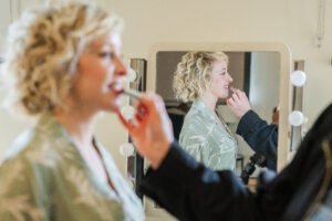 A woman with curly blonde hair is getting her lips applied with lipstick by another person. She is reflected in a mirror behind her, which has round lights on its sides. The woman is wearing a green, patterned, open-collared top.