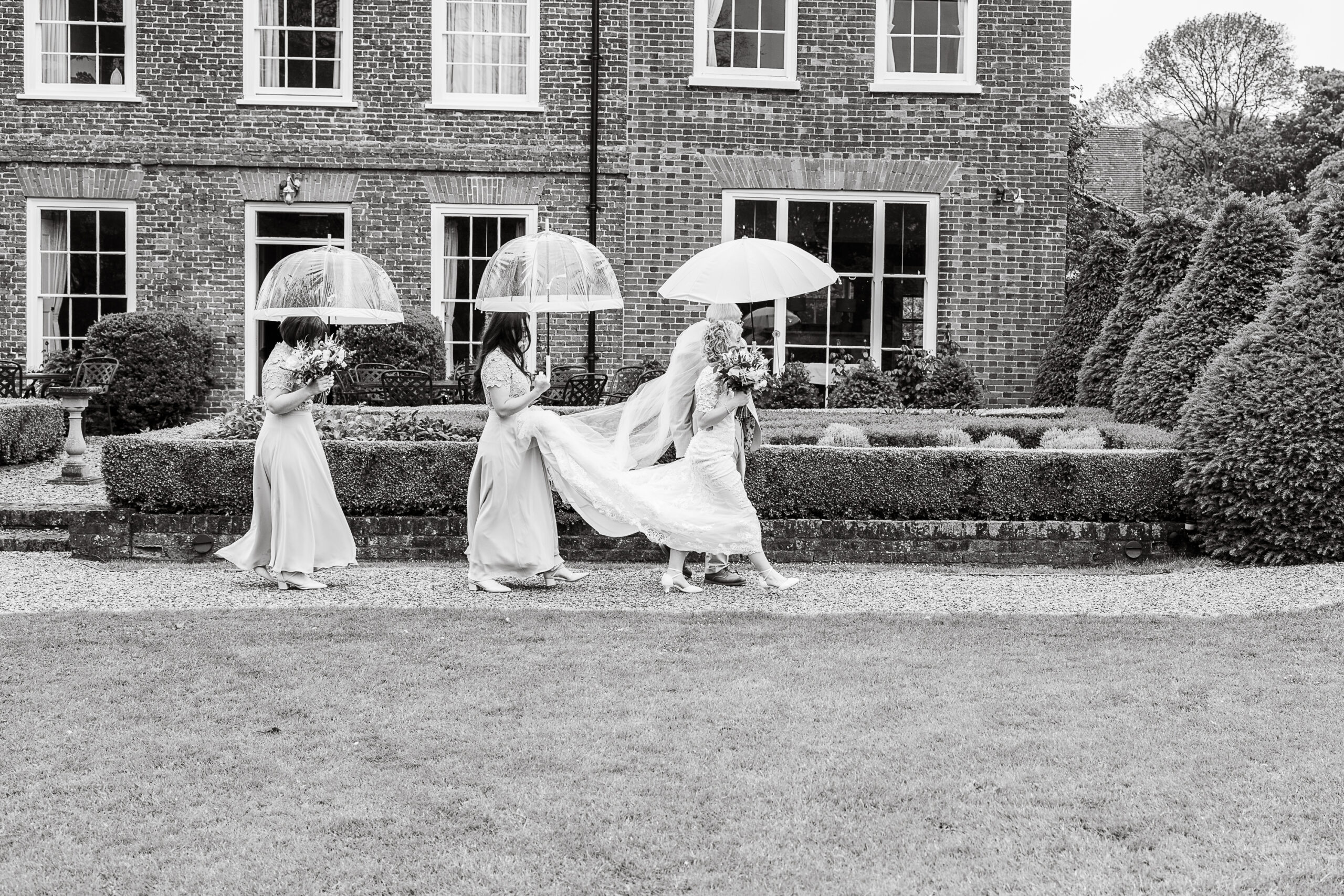 A black-and-white photo shows three bridesmaids and a bride walking in front of a large brick building. The bridesmaids carry flower bouquets and each holds a clear umbrella while one helps lift the bride's dress train as they move forward.