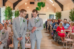 Two grooms in light gray suits and green ties stand at the front of a room filled with seated guests. One groom smiles while checking his watch as the other looks at him. The room is decorated with greenery and hanging plants, and people look on expectantly.