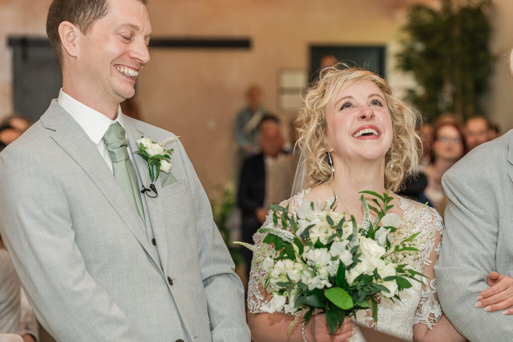 A joyful couple is smiling widely during their wedding ceremony. The bride is holding a bouquet of white flowers and greenery, wearing an elegant lace dress. The groom is dressed in a light grey suit with a mint green tie. They are surrounded by guests in the background.