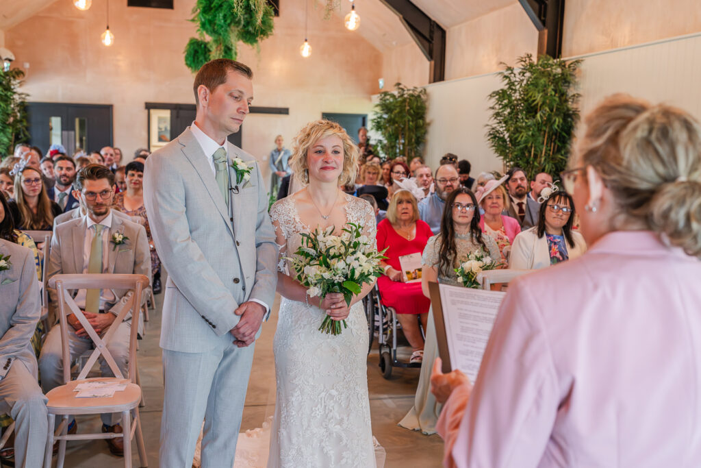 A bride and groom stand at the altar during their wedding ceremony, with the officiant reading from a script. The bride holds a bouquet of white flowers, and the groom stands beside her in a light gray suit. Guests watch from their seats in a warmly lit, decorated venue.