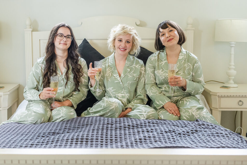 Three women in matching green pajamas with white leaf designs sit on a bed, smiling and holding champagne glasses. They appear to be enjoying a relaxed moment together. The bed has white linens and a black and white patterned blanket at the foot.