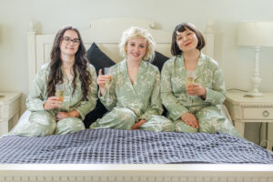 Three women in matching green pajamas with white leaf designs sit on a bed, smiling and holding champagne glasses. They appear to be enjoying a relaxed moment together. The bed has white linens and a black and white patterned blanket at the foot.