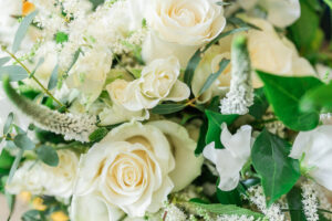 A close-up of a flower arrangement featuring a variety of white flowers, primarily roses, mixed with lush green leaves and white accent flowers. The bouquet appears fresh and delicate, showcasing the intricate details of the petals and foliage.