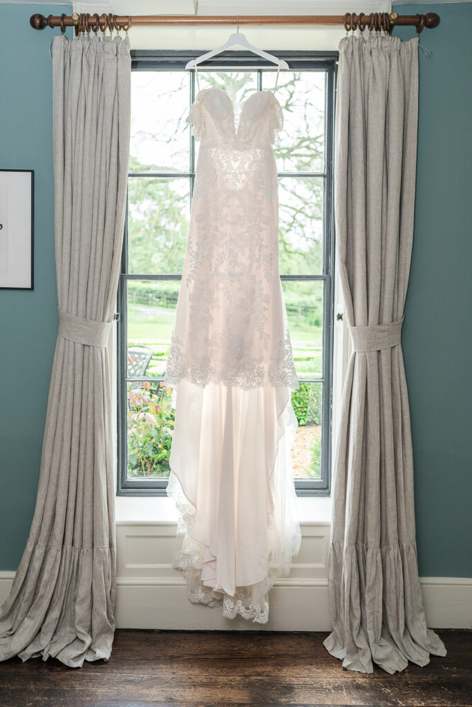 A sleeveless, intricately lace-embellished wedding dress hangs on a hanger in front of a large window framed by light gray curtains. The dress features a flowing train and delicate floral patterns. The bright outdoor scenery is visible through the window behind it.