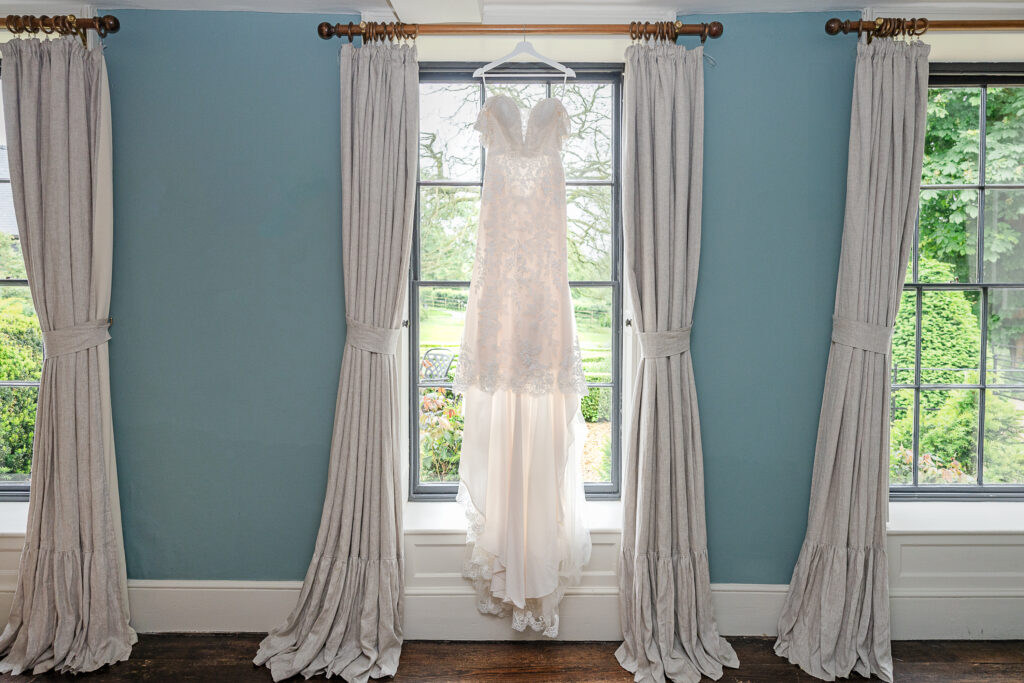 An elegant wedding dress is hanging on a hanger in front of a large window flanked by two grey curtains. The dress is white with lace detailing and a flowing train. Through the window, a lush green garden can be seen.