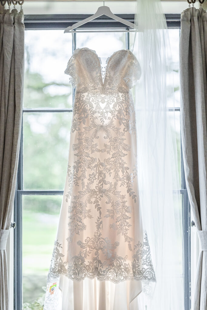 A white lace wedding dress with an intricate floral pattern hangs on a hanger in front of a large window. The dress has off-the-shoulder sleeves and a flowing veil drapes beside it. The window is framed by light gray curtains.