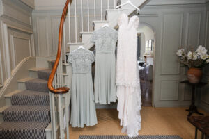 Three dresses hang from hangers on a staircase railing. One is a white lace wedding dress, and the other two are light gray bridesmaid dresses with lace tops. A vase with white flowers is on a wooden table nearby. The background features beige paneled walls and a carpeted staircase.