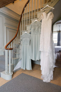 Three sage green bridesmaid dresses and one white lace wedding gown hang on the railing of a gray staircase with wooden handrails. The dresses are displayed in a well-lit room with a striped rug on the floor and a table partially visible in the background.