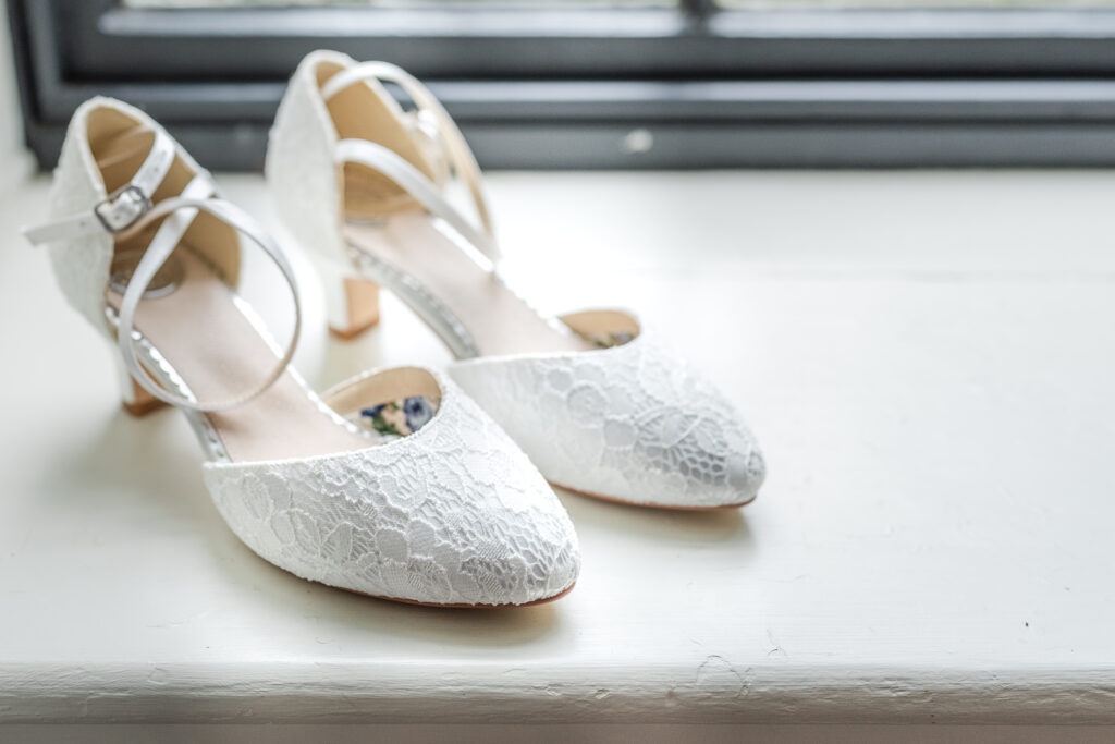 A pair of white lace wedding shoes with chunky heels and ankle straps are placed on a white windowsill. Soft natural light filters through the window, highlighting the delicate lace details on the shoes.