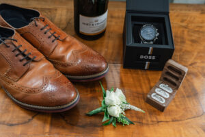 A pair of polished brown leather brogues, a boutonnière with white flowers, a wooden box with three silver rings, a black boxed wristwatch, and a bottle of Moët & Chandon champagne are displayed on a wooden surface.