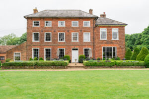 An elegant, large, three-story red-brick mansion with symmetrical windows and a central white door. The front facade is complemented by neatly trimmed hedges, manicured gardens, and a green lawn, set against a backdrop of lush trees under an overcast sky.