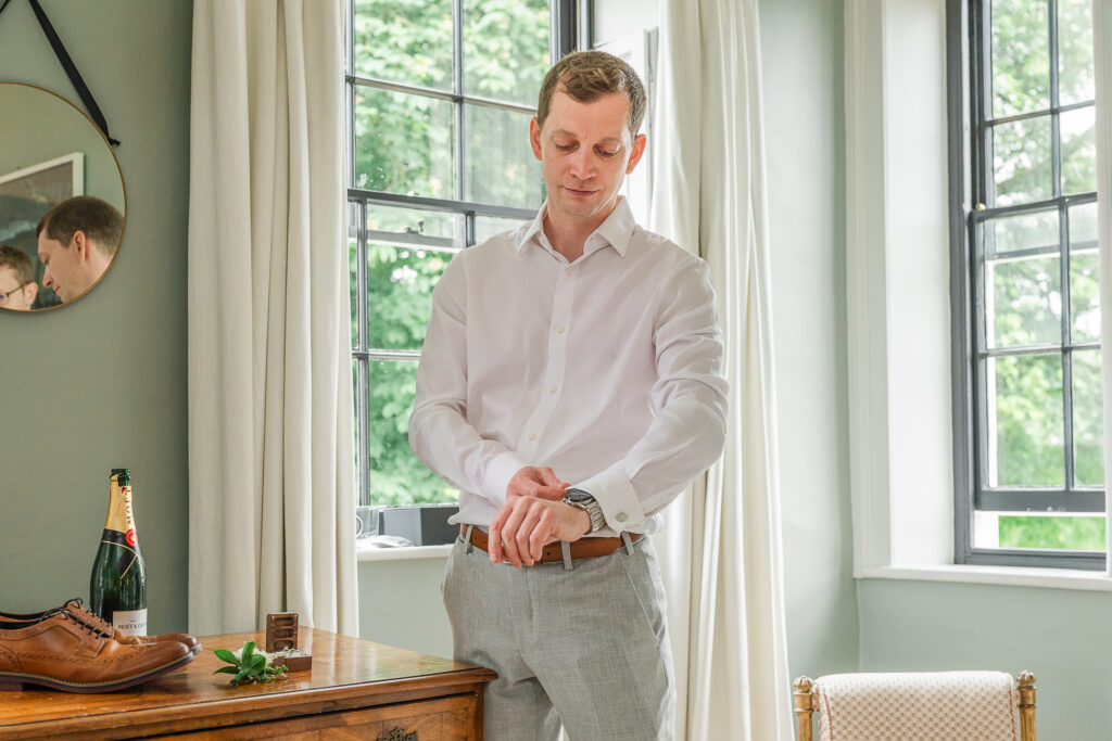 A man stands in a well-lit room with large windows, wearing a white shirt and gray trousers. He adjusts his wristwatch by a wooden table that holds a pair of brown shoes, a bottle of champagne, and a small box. A mirror reflecting part of the room hangs on the wall.