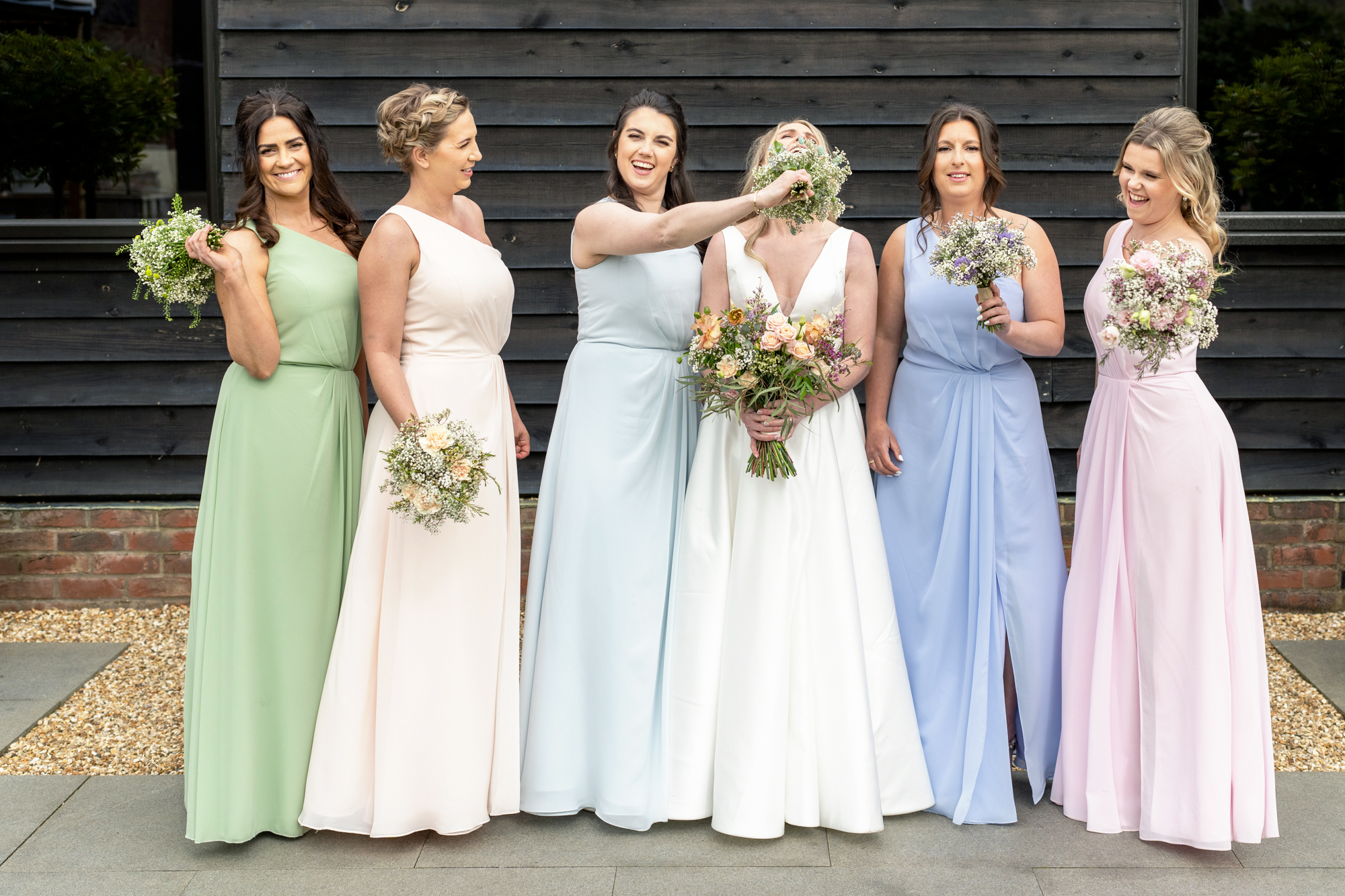 A bride in a white dress stands with five bridesmaids, who are wearing dresses in pastel colors, in front of a dark wooden wall at Milling Barn, hertfordshire. One bridesmaid playfully holds a bouquet of flowers in front of the bride’s face, causing both to laugh.