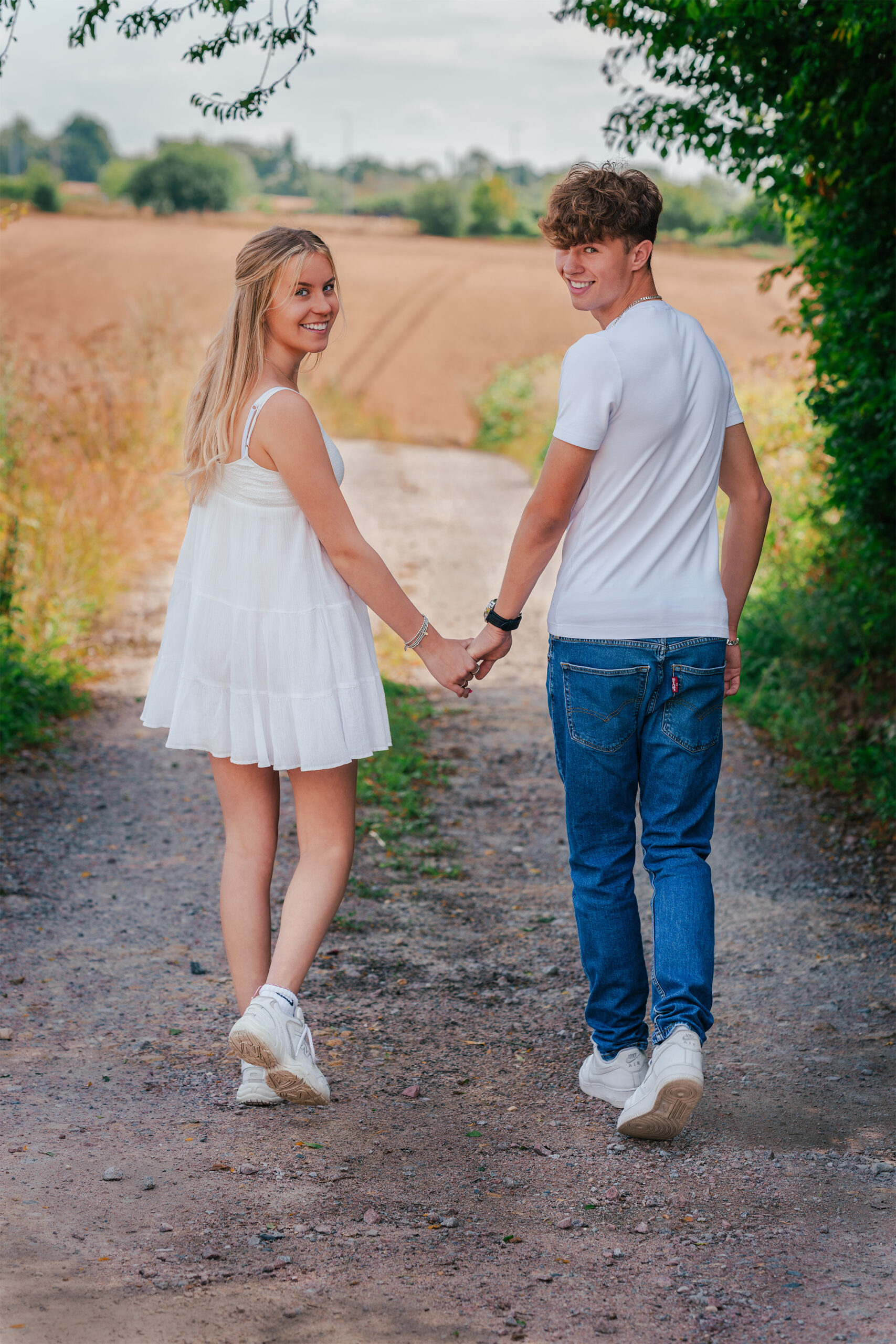 A young couple holds hands while walking down a rustic path surrounded by greenery and fields. Both are smiling and looking back towards the camera. The girl wears a white dress and sneakers, and the boy wears a white t-shirt, jeans, and sneakers.