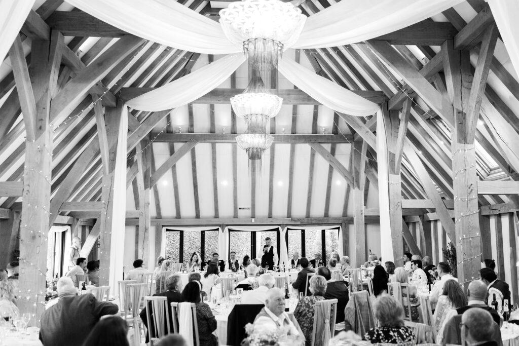 A black and white photograph capturing a wedding reception Old Kent Barn. Guests are seated at round tables, facing a couple standing at the head table in the distance. Chandeliers add elegance to the setting.