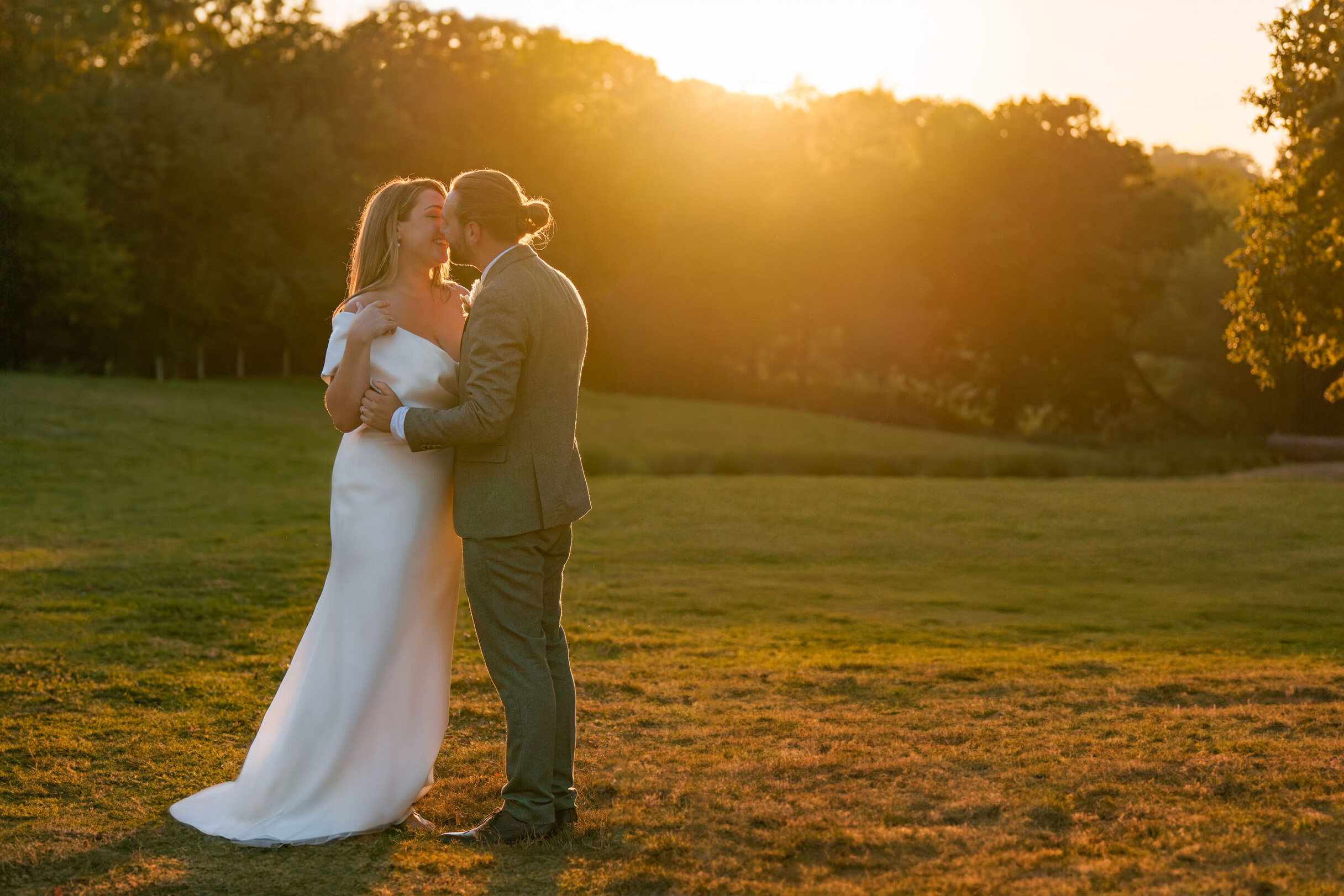 A bride and groom stand in a grassy field with trees and a golden sunset in the background at Old Oak Barn, Frame Farm, Kent. The bride wears a flowing white dress and the man is in a green suit. They are holding each other closely and gazing into each other’s eyes, creating a romantic moment.