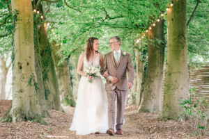 A bride in a flowing white dress walks arm-in-arm with her father in a checkered suit down a tree-lined path adorned with hanging lights. Both are smiling cheerfully, surrounded by the green foliage and dappled sunlight. The bride holds a bouquet of flowers.