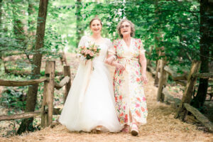 A bride in a flowing white dress holds a bouquet as she walks arm-in-arm with her mother in a floral dress. They are outdoors on a straw-covered path surrounded by trees and wooden fencing, both smiling and looking joyful at the Paper Mill, Kent