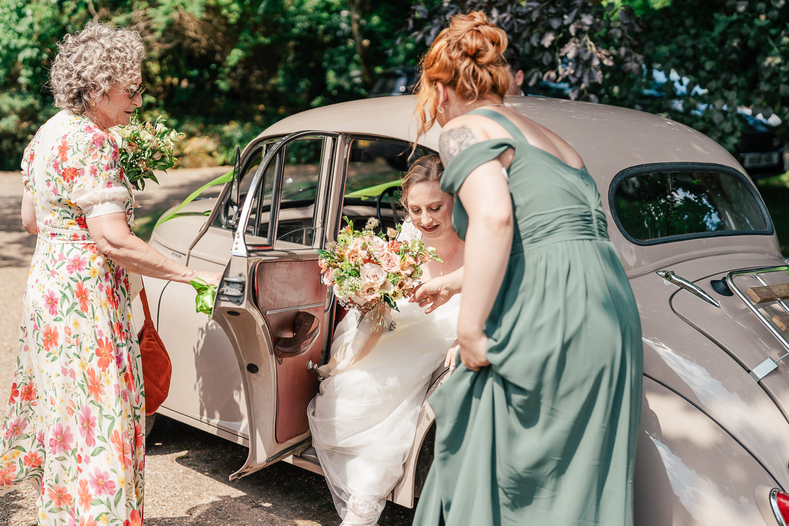 A bride in a white gown steps out of a vintage beige car, assisted by a woman in a green dress. Her mother in a floral dress stands nearby holding her bouquet. The scene is set outdoors on a sunny day, surrounded by lush greenery at the Paper Mill, Kent