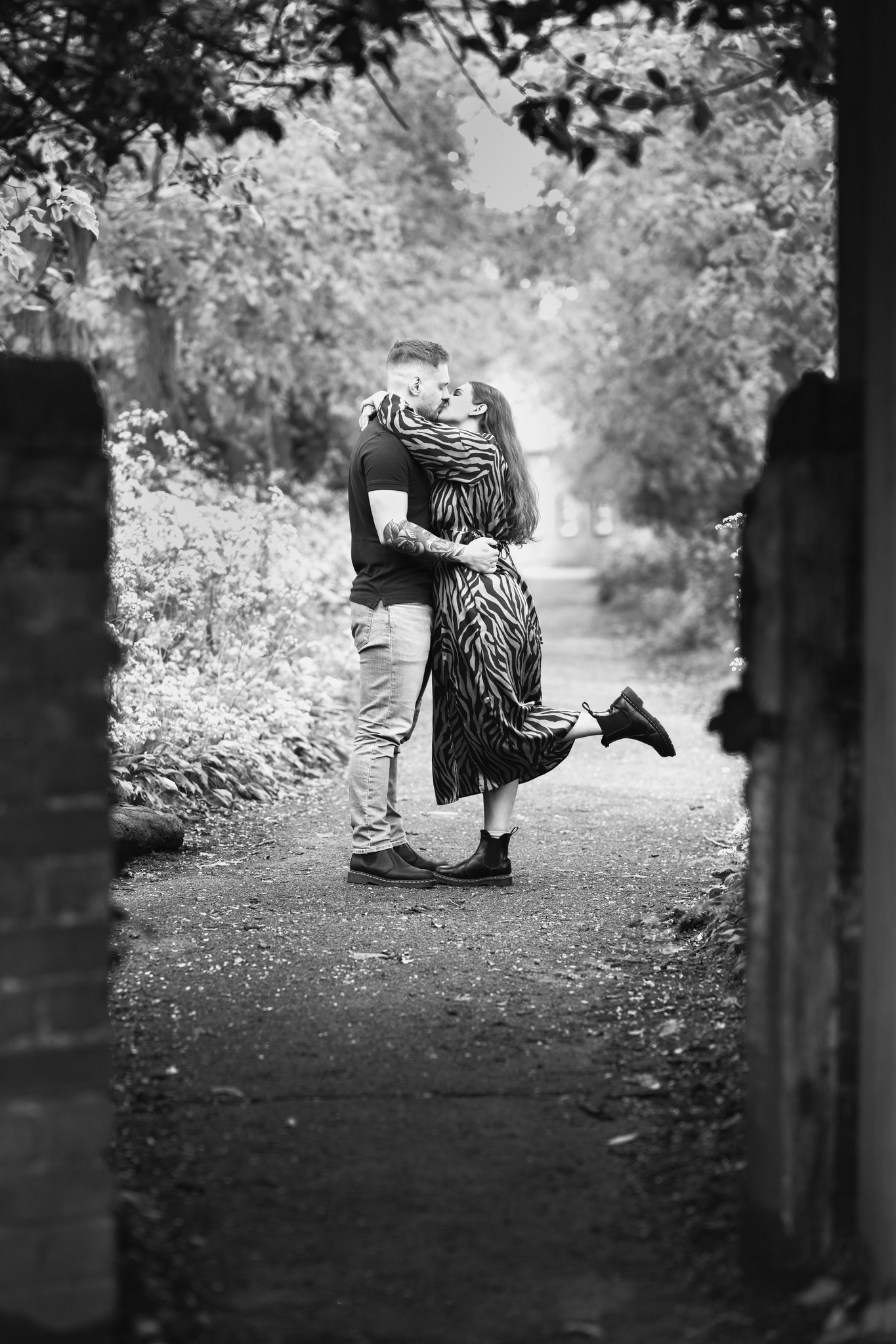 A couple embraces and shares a kiss in a leafy, tree-lined pathway. The woman, dressed in a long patterned dress, lifts one leg in the air, while the man, in a t-shirt and jeans, wraps his arms around her. The scene is framed by a partially open gate.