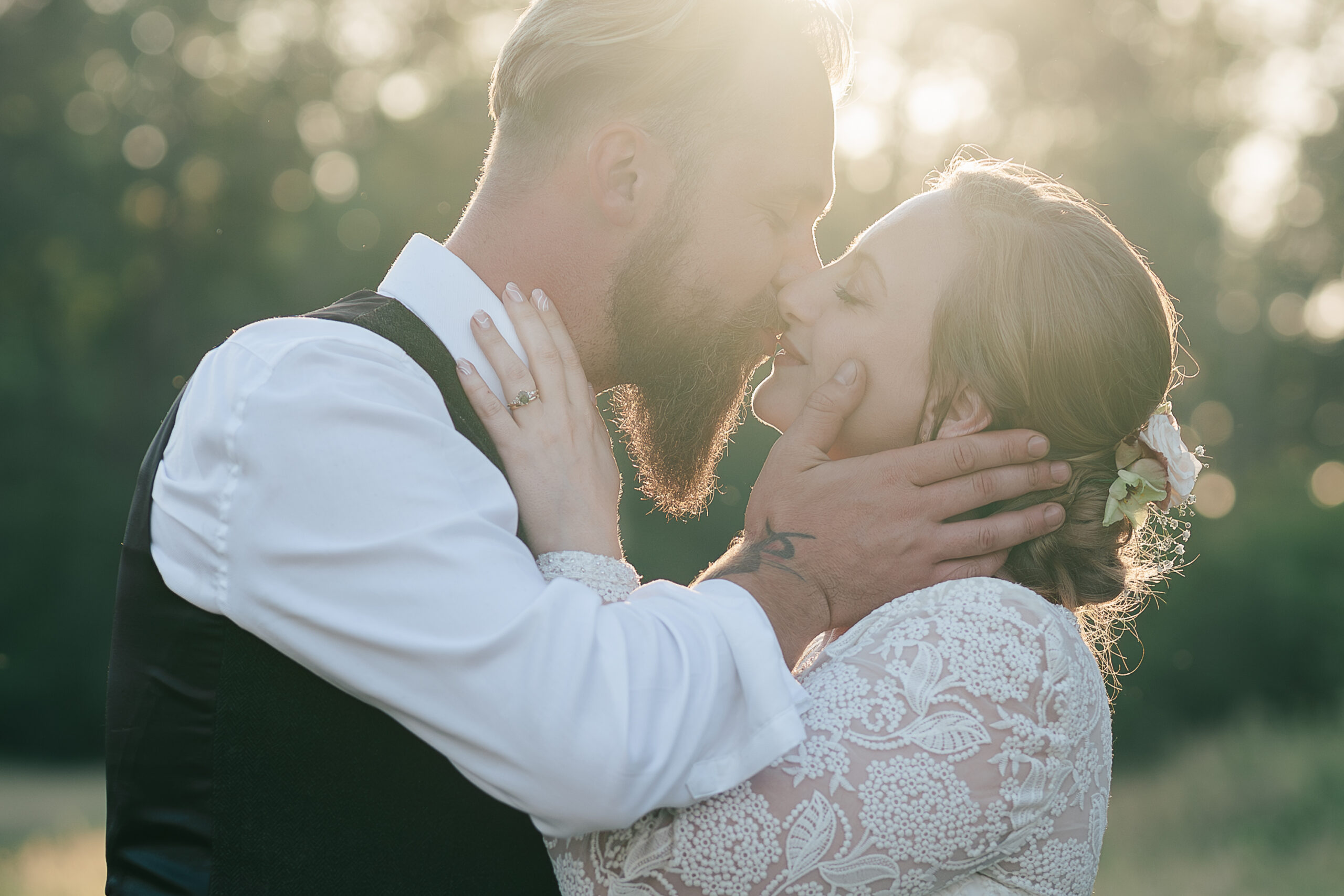 A bride and groom, share a tender kiss outdoors at the Paper Mill wedding venue, Kent, captured beautifully by a Hertfordshire Wedding Photographer. The groom, with a beard and blonde hair, gently holds the bride's face. The bride, wearing a lace dress with flowers in her hair, smiles with her eyes closed as sunlight filters through trees in the background.