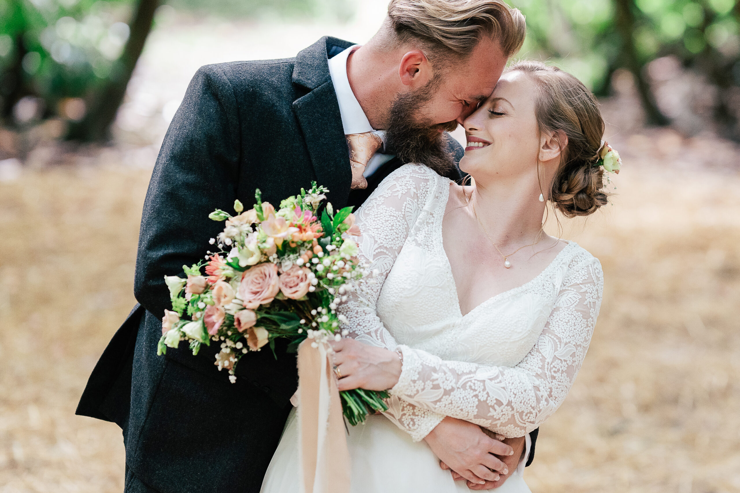 A bride in a white lace gown and a groom in a dark suit share an intimate, joyful moment outdoors. The bride holds a bouquet of flowers, and the groom leans in close, nuzzling her forehead and making her smile. The background shows greenery and a wooded area.