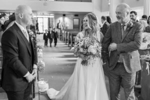 A bride holding a bouquet of flowers smiles as she stands next to an elderly man, presumably her father, who is holding her arm. They face a man in a suit, likely the groom. People are seated in the background of the church, and there are flower decorations along the aisle.