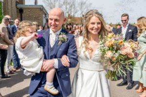 A smiling bride and groom walk out of a building, holding a child in their arms. The groom wears a navy blue suit and the bride a white dress, holding a bouquet of flowers. Confetti is being thrown by the surrounding crowd, visible in the background.