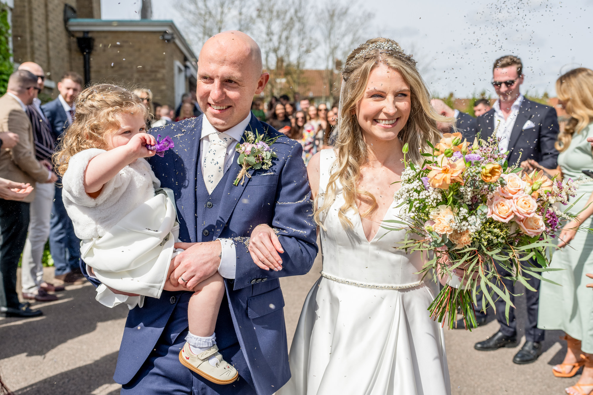 A smiling bride and groom walk out of a building, holding a child in their arms. The groom wears a navy blue suit and the bride a white dress, holding a bouquet of flowers. Confetti is being thrown by the surrounding crowd, visible in the background.