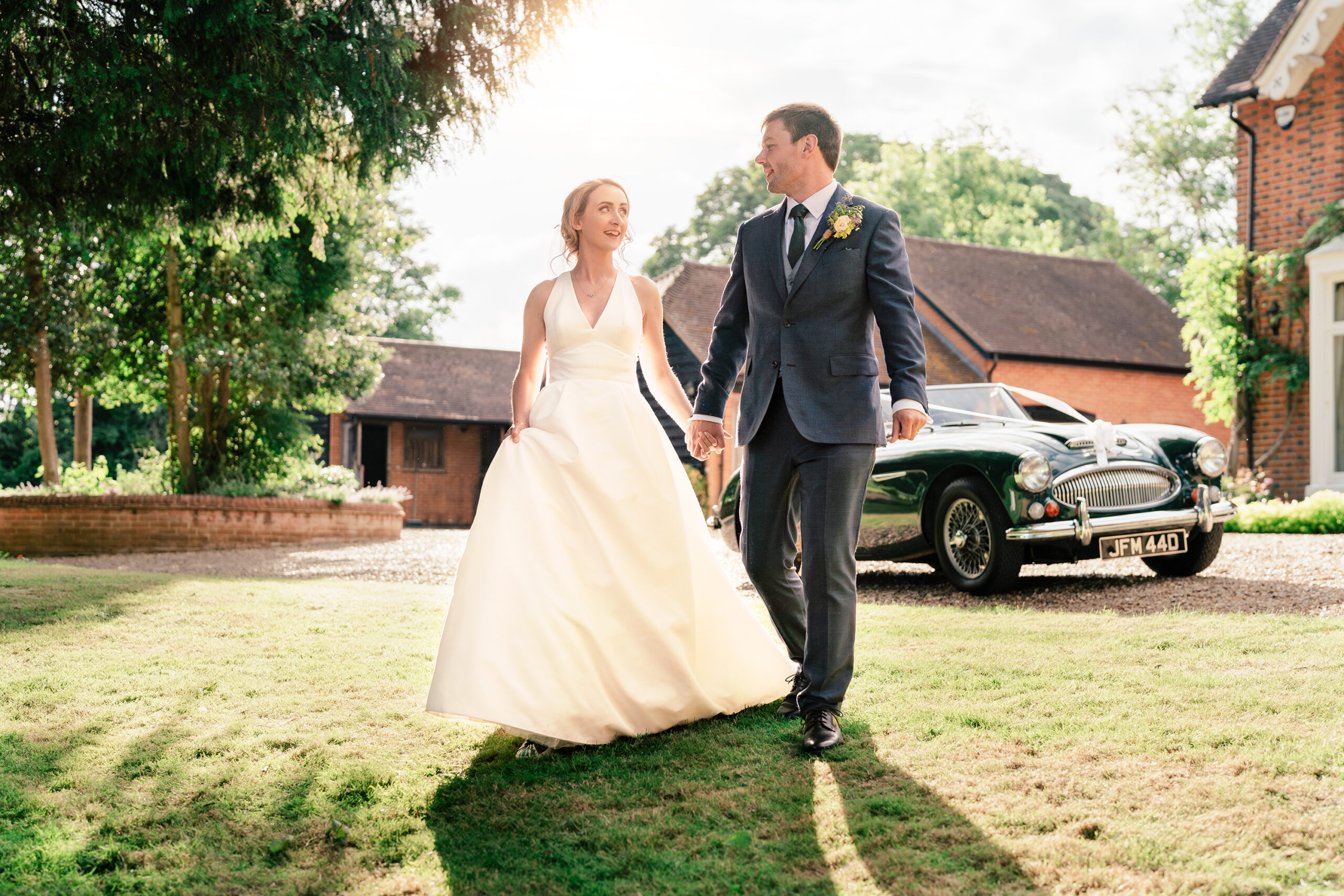 A bride in a white gown and a groom in a dark suit walk hand in hand on a grassy lawn. They are near a classic green car parked on a gravel driveway, with trees and stables in the background. The sun is shining, casting a warm glow on the scene.