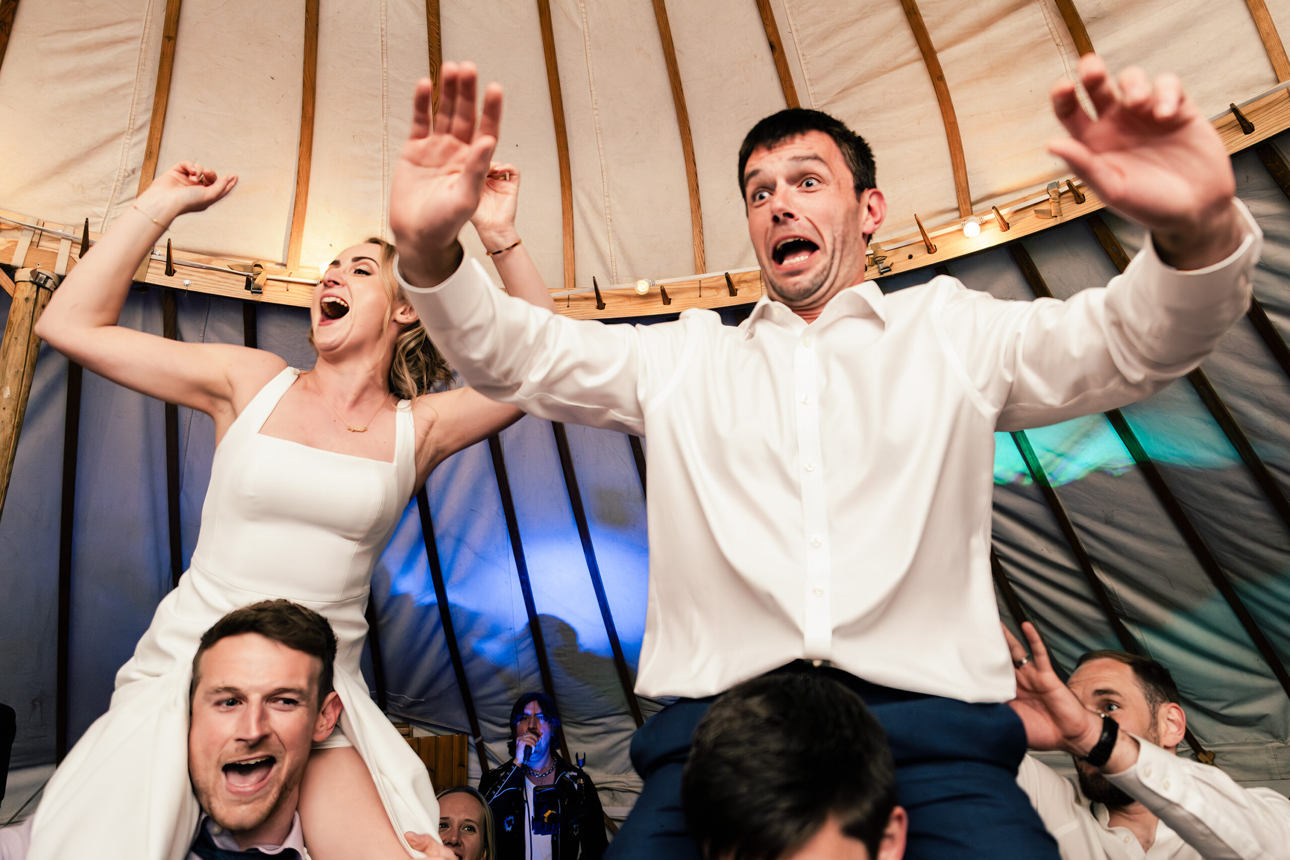 A joyful bride and a groom are lifted on someone's shoulders during a lively wedding celebration inside a tent. They are both smiling and raising their hands in excitement. The bride wears a white dress, and the groom is in a white shirt. Guests are around them.