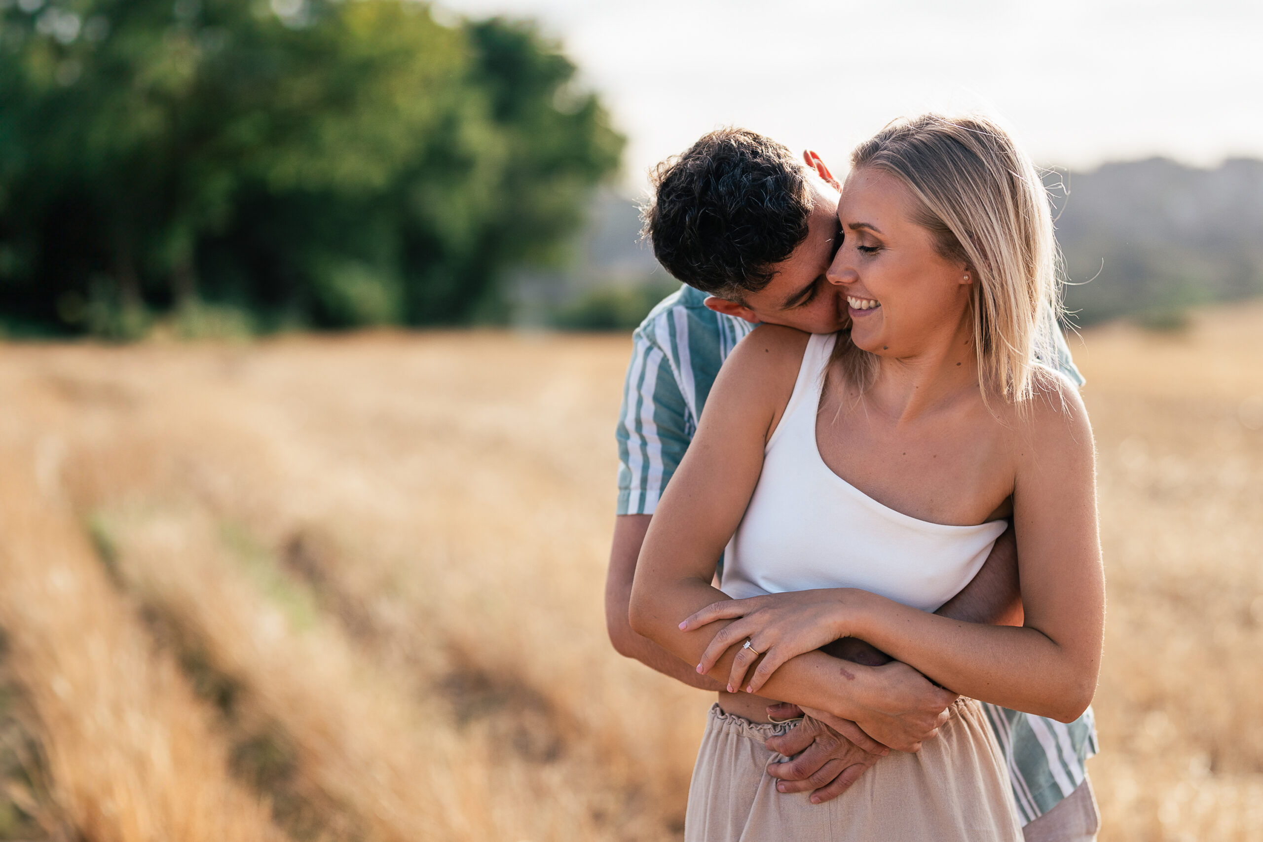 A man and woman embrace in a wheat field. The woman is smiling and looking to the side, while the man stands behind her with his arms around her waist and his face close to hers. Both are dressed in light, summer clothes, and the field extends into the background. denhelderweddings.com