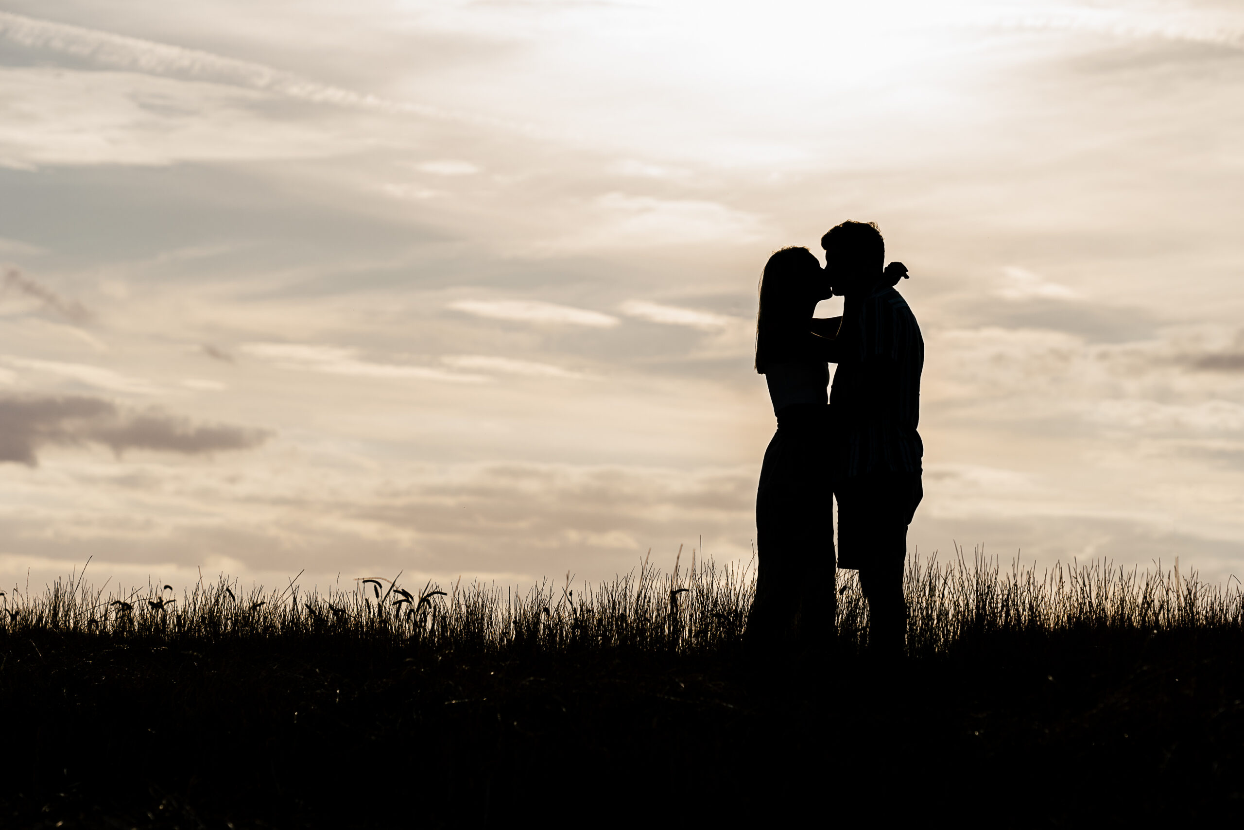 Silhouette of a couple embracing with a kiss in a field against a backdrop of a bright, cloudy sky. The sun appears low, creating a serene and romantic atmosphere.