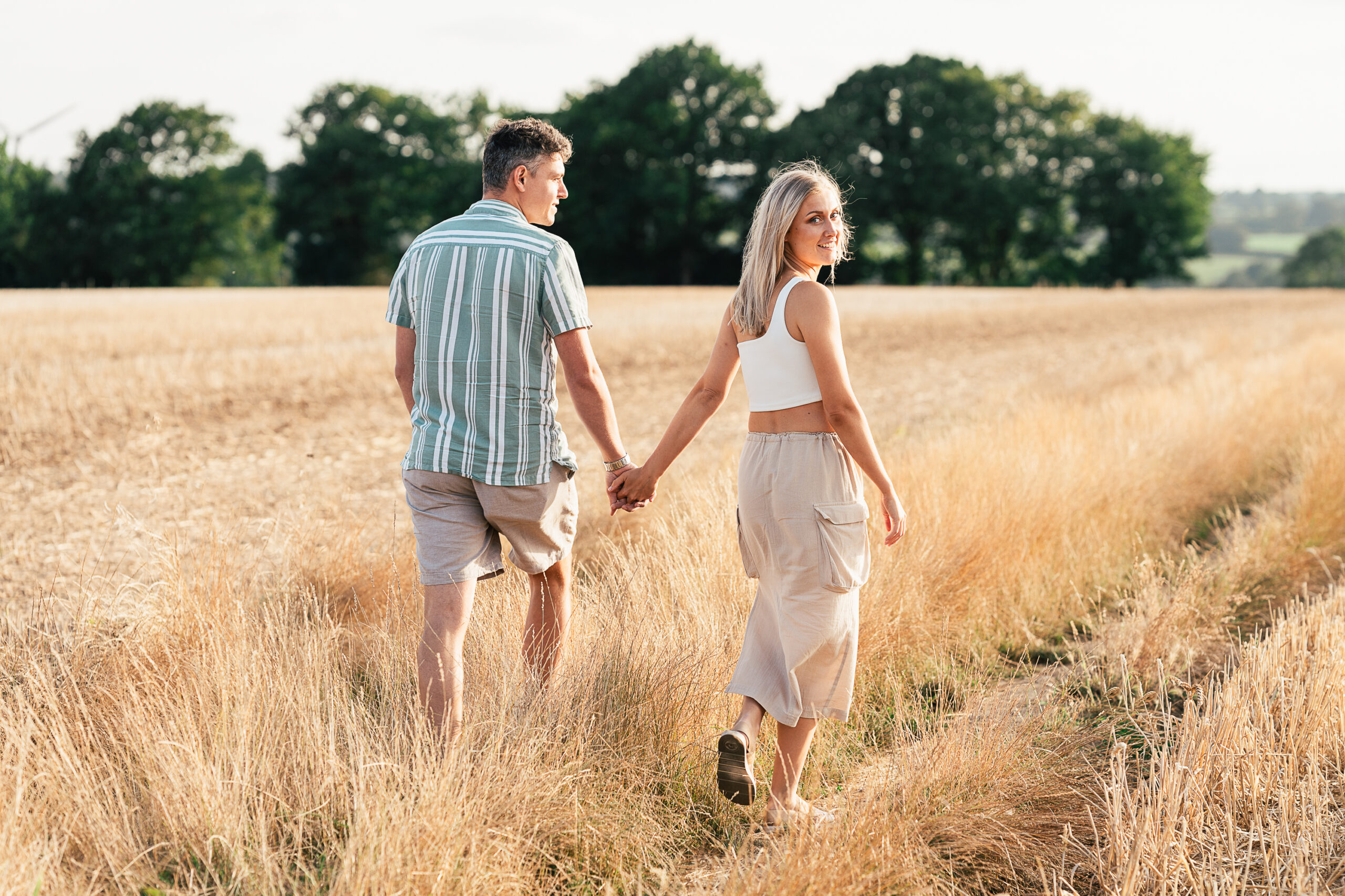 A couple walks hand in hand through a field of tall, golden grass. The woman glances back with a smile, wearing a white top and beige skirt, while the man wears a striped shirt and shorts. Trees and distant hills are visible under a partly cloudy sky.