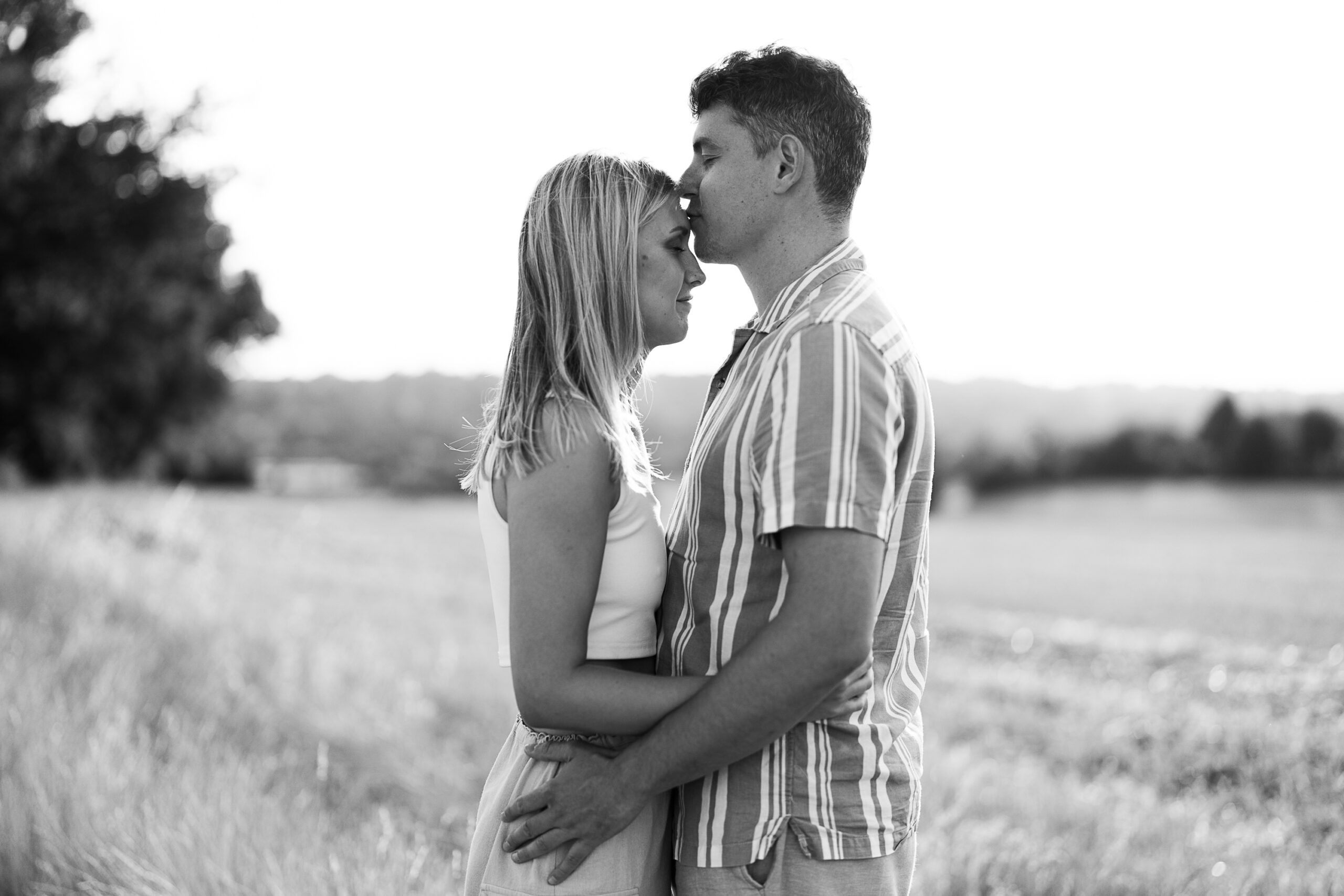 A black and white photo of a couple standing in an open field. The man is giving the woman a forehead kiss while they embrace each other. They're both dressed in casual summer clothes, and the background features trees and a distant horizon under bright daylight.