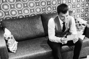 A man dressed in formal attire, including a vest and tie, sits on a couch with patterned pillows. He is smiling and reading a card or letter. The wall behind him features a circular, geometric pattern. The scene is captured in black and white.