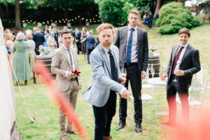 Four men dressed in suits stand on a grassy lawn at an outdoor event. One man in a light blue jacket is throwing an object, while the others watch. There are lights strung in the background and tables with floral arrangements nearby.