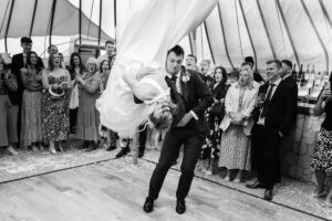 A man in a suit, holding a woman in a wedding dress as she performs an impressive dance lift, surrounded by delighted onlookers inside a tent. Confetti is scattered on the floor, and guests in the background are clapping and smiling.