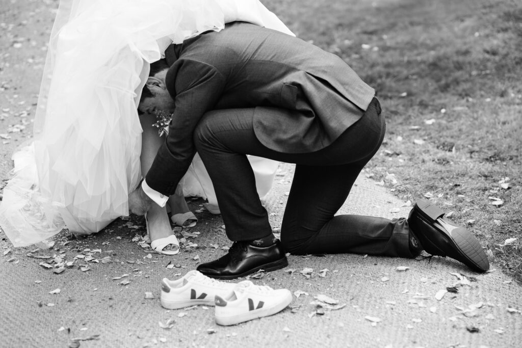 A black-and-white photo of a groom, wearing a suit and kneeling on the ground, helping his bride adjust or remove her shoe. The bride's dress and veil partially cover her face. A pair of sneakers are placed on the ground beside them.